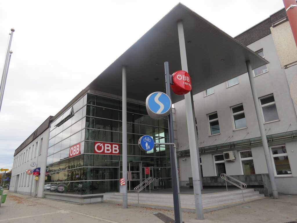 Schrägansicht der verglasten Bahnhofshalle von St. Valentin mit großem Flugdach und ÖBB-Logos, rechts der helle mehrgeschoßige Altbau, davor Stufen mit Geländern und ein S-Bahn-Schild unter bewölktem Himmel