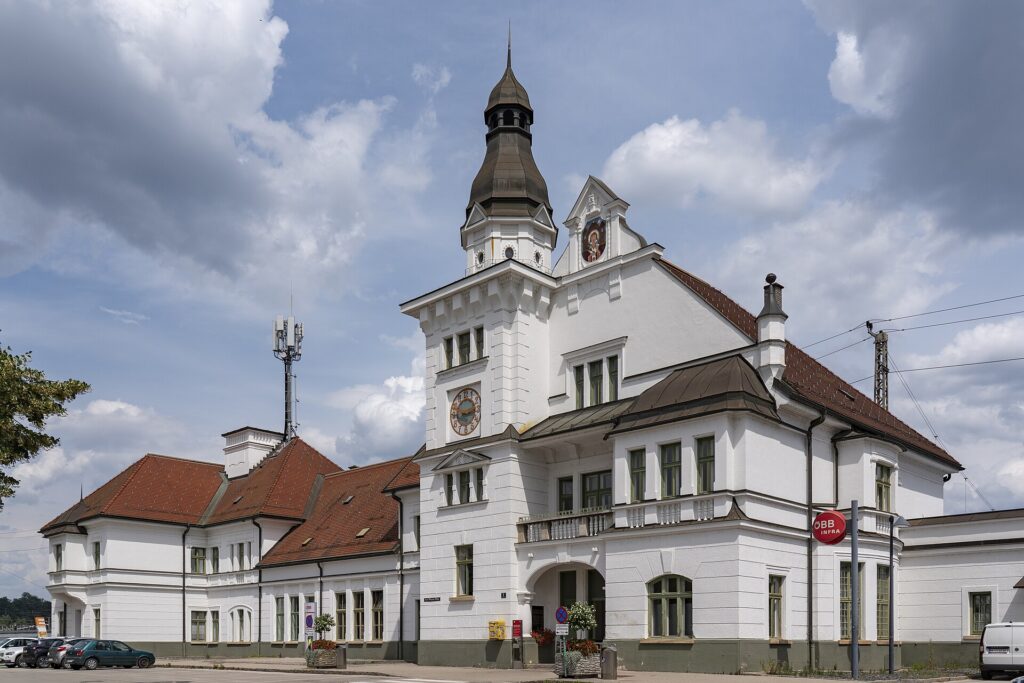 Schrägansicht des weißen Aufnahmsgebäudes von St. Veit an der Glan mit Uhrturm und Laternenaufsatz, roten Ziegeldächern und Rundbogeneingang, davor geparkte Autos unter bewölktem Himmel