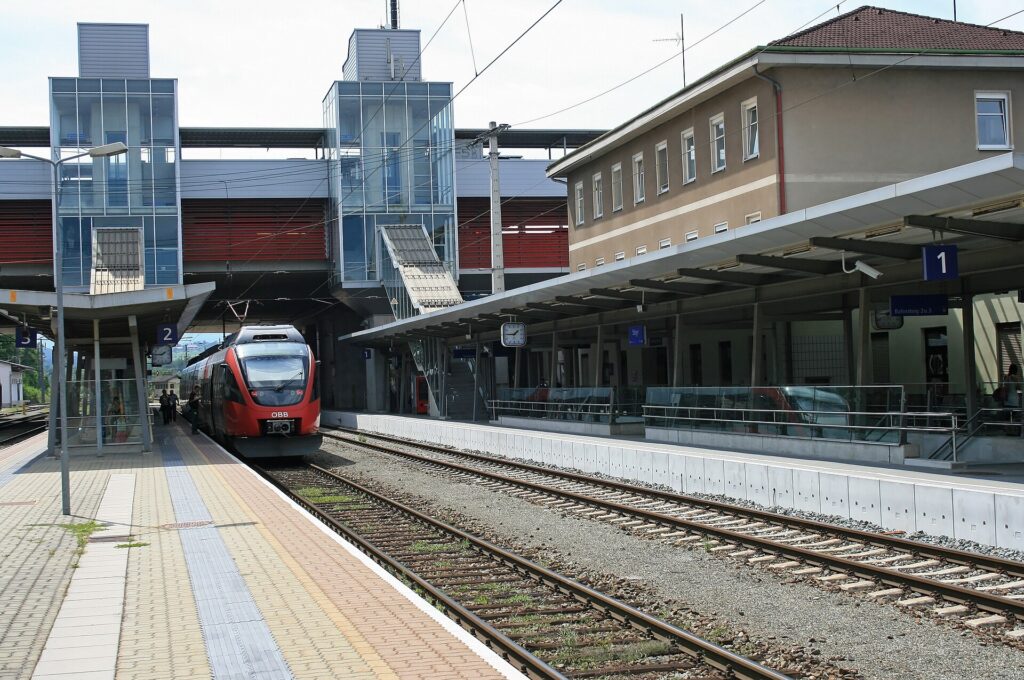Blick vom Mittelbahnsteig in Steyr auf den überdachten Bahnsteig 1 und das zweistöckige Bahnhofsgebäude rechts, links die verglasten Stiegen- und Lifttürme vor dem Parkdeck über den Gleisen, darunter ein roter Nahverkehrstriebwagen
