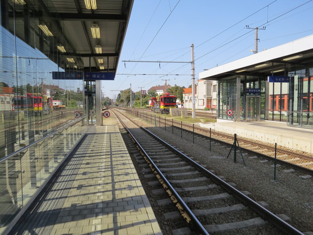 Überdachter Bahnsteig mit gläserner Windschutzwand am Bahnhof Stockerau, daneben die Gleise und gegenüber ein moderner Bahnsteig mit verglaster Wartehalle unter blauem Himmel.