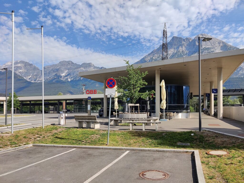 Ein flaches Betondach auf runden Stützen über einem verglasten, gerundeten Pavillon mit Café-Tischen, davor Sitzbänke, ein Baum und eine Bushaltestelle, links das ÖBB-Logo, im Hintergrund Kalkberge unter teils bewölktem Himmel