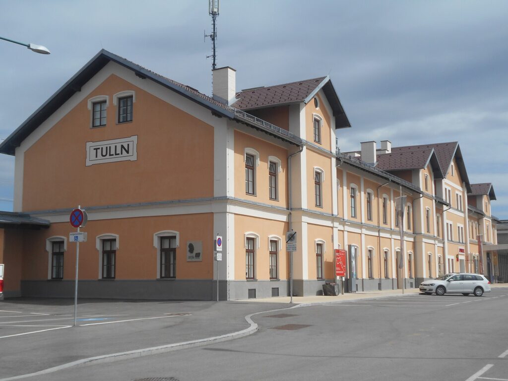 Das renovierte zweigeschoßige, ockerfarbene Bahnhofsgebäude von Tulln mit weißen Fensterumrahmungen, braunem Dach und einem Giebel mit dem Schriftzug TULLN, davor ein leerer Vorplatz mit Parkmarkierungen, bewölkter Himmel