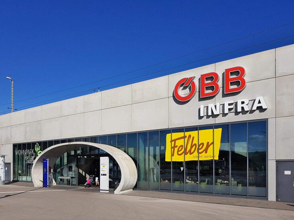 Die flache Betonfassade der Bahnhofshalle Tullnerfeld mit Glasfront und einem geschwungenen Betonbogen über dem Eingang, darüber die Oberleitung, tiefblauer Himmel; am Eingang kleine Personen mit Kinderwagen