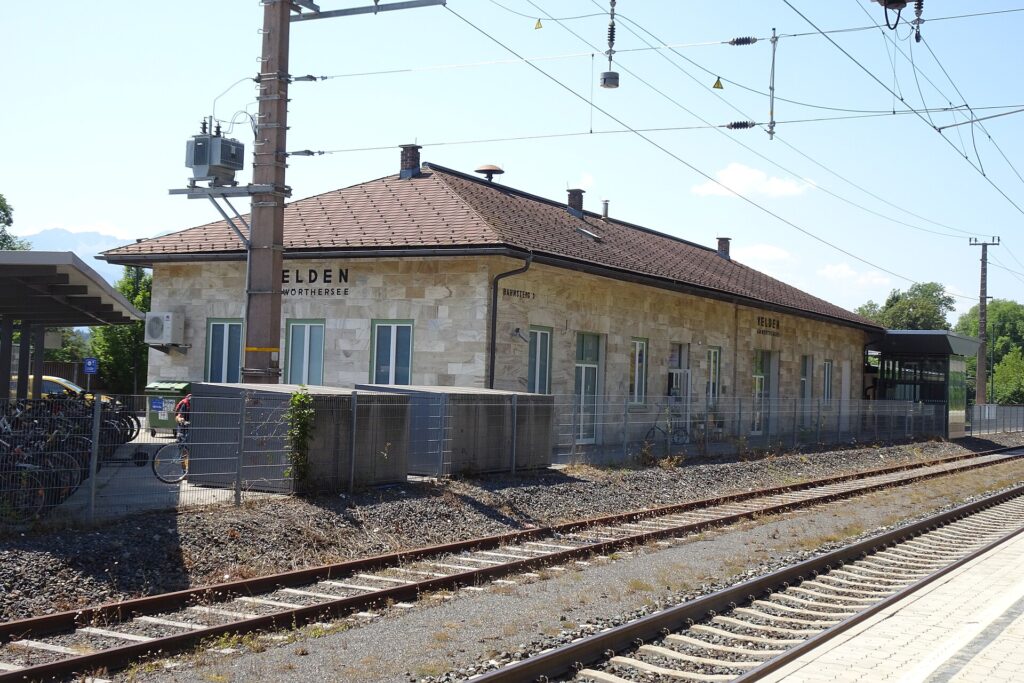 Das lang gestreckte, eingeschoßige Aufnahmsgebäude von Velden aus hellen Steinquadern mit braunem Walmdach und dem Schriftzug Velden am Wörthersee, davor Gleise und die Bahnsteigkante, links eine überdachte Fahrradabstellanlage