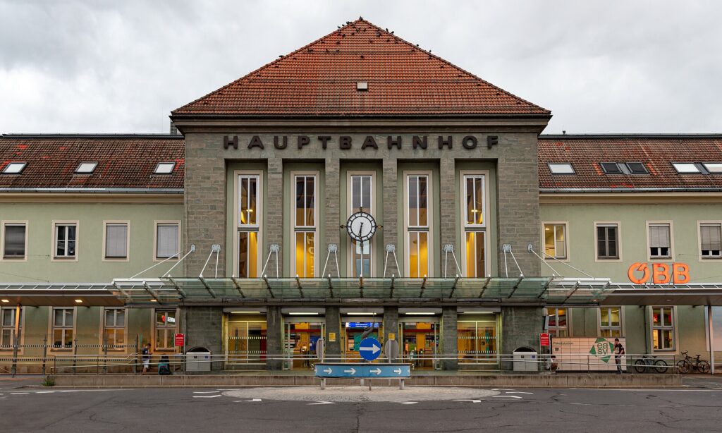 Frontalansicht des Villacher Hauptbahnhofs: ein hoher Mittelbau aus grauem Stein mit Walmdach, dem Schriftzug HAUPTBAHNHOF, hohen Fenstern und einer Uhr, davor ein gläsernes Vordach über dem Eingang; links und rechts die hellgrünen Seitenflügel, rechts das ÖBB-Logo
