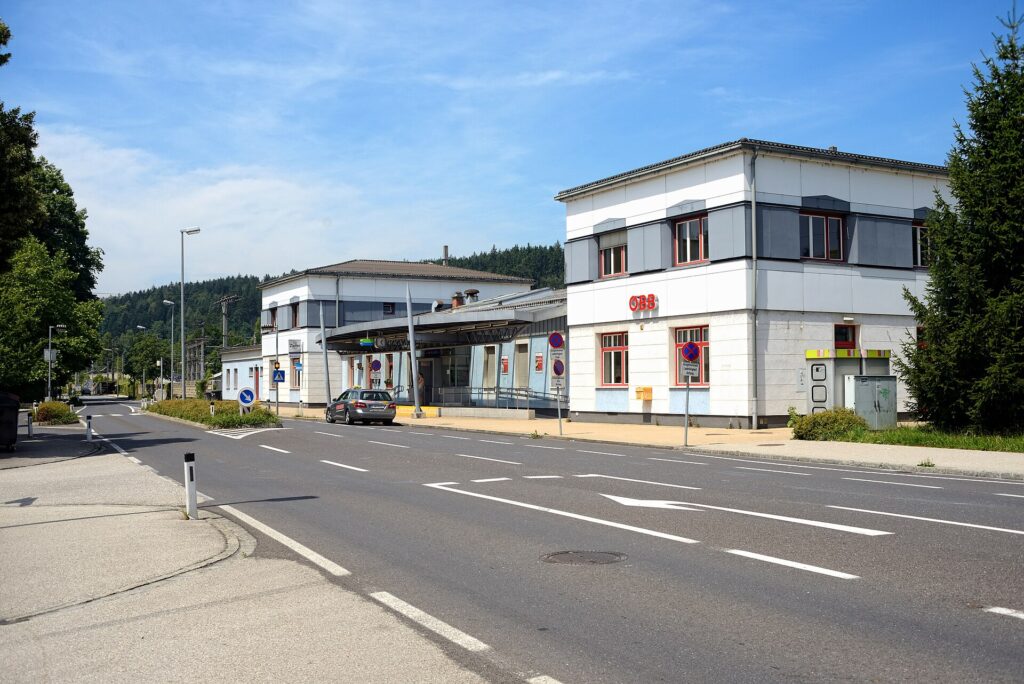 Straßenansicht des hellen, zweigeschoßigen Aufnahmsgebäudes von Vöcklabruck mit ÖBB-Logo und überdachtem Eingangsbereich an der Bahnhofstraße unter blauem Himmel