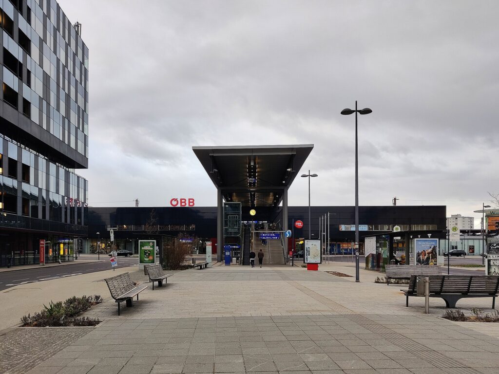 Blick über den gepflasterten Bahnhofplatz auf das flache, dunkle Bahnhofsgebäude mit ÖBB-Schriftzug; in der Mitte ein großes Vordach über einer Treppe und Rolltreppe hinauf zum Steg, links ein Bürogebäude mit Glasfassade, rechts Wartehäuschen der Bushaltestelle