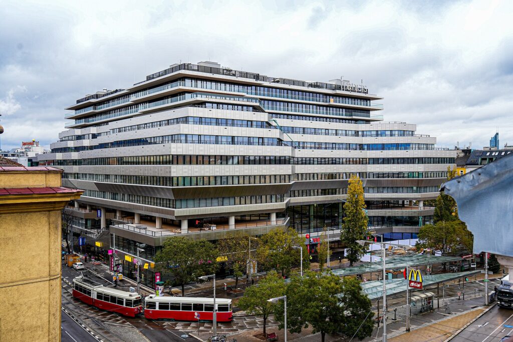 Der helle, mehrgeschoßige Bürobau „Francis“ mit Bandfassade über dem Bahnhof, davor der Julius-Tandler-Platz mit zwei roten Straßenbahnen, Haltestellendach und Bäumen unter bewölktem Himmel
