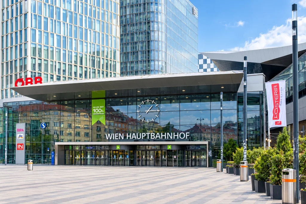 Haupteingang des Wiener Hauptbahnhofs am Südtiroler Platz: Glasfassade mit großer Uhr und dem Schriftzug Wien Hauptbahnhof unter einem weit auskragenden Vordach, links das rote ÖBB-Logo, dahinter Bürotürme, davor der gepflasterte Vorplatz mit Pflanzentrögen (KI-Nachbildung nach einem Foto)