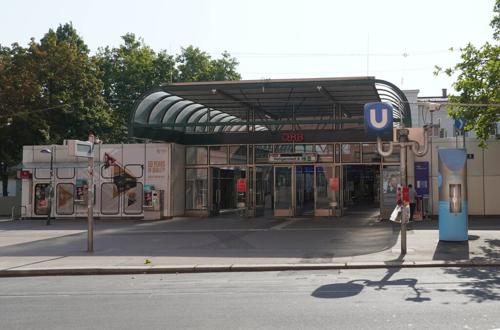 Der Eingang des Bahnhofs Heiligenstadt mit geschwungenem, verglastem Vordach, rotem ÖBB-Schriftzug und U-Bahn-Schild, davor Gehsteig, Straße und ein blauer Trinkwasserbrunnen, im Hintergrund Bäume