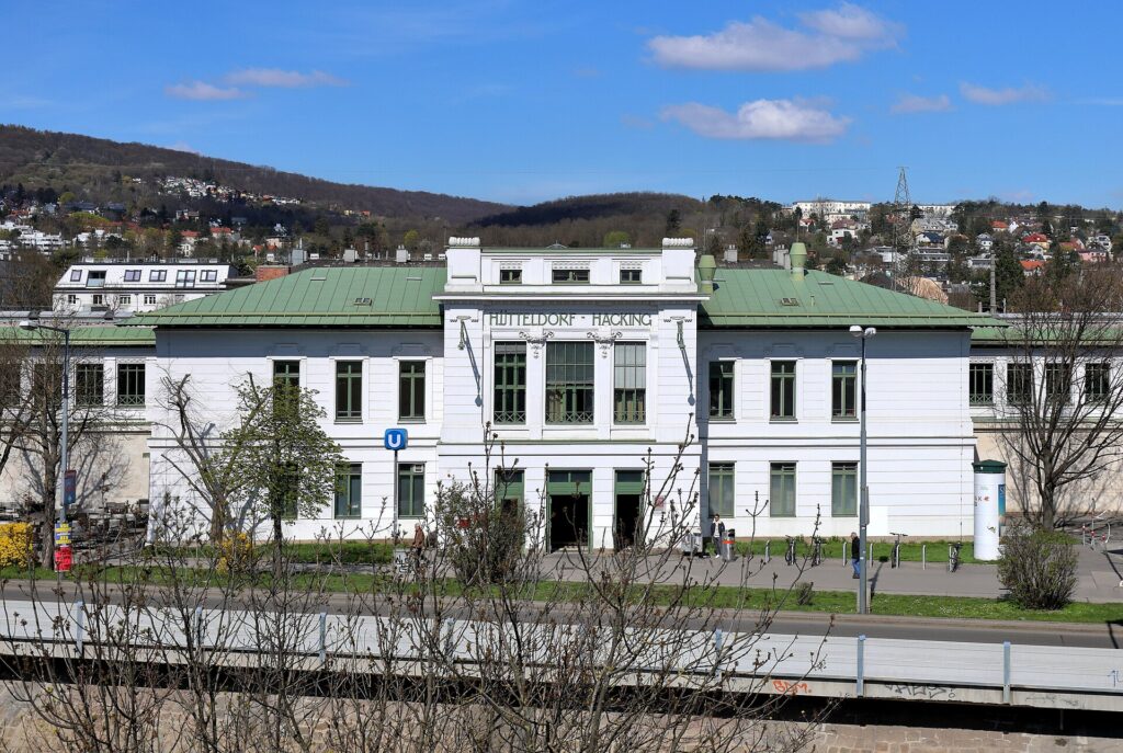Das weiße südliche Aufnahmsgebäude des Bahnhofs Hütteldorf mit grünem Dach und dem Schriftzug Hütteldorf-Hacking, aufgenommen vom anderen Ufer des Wienflusses an einem sonnigen Tag
