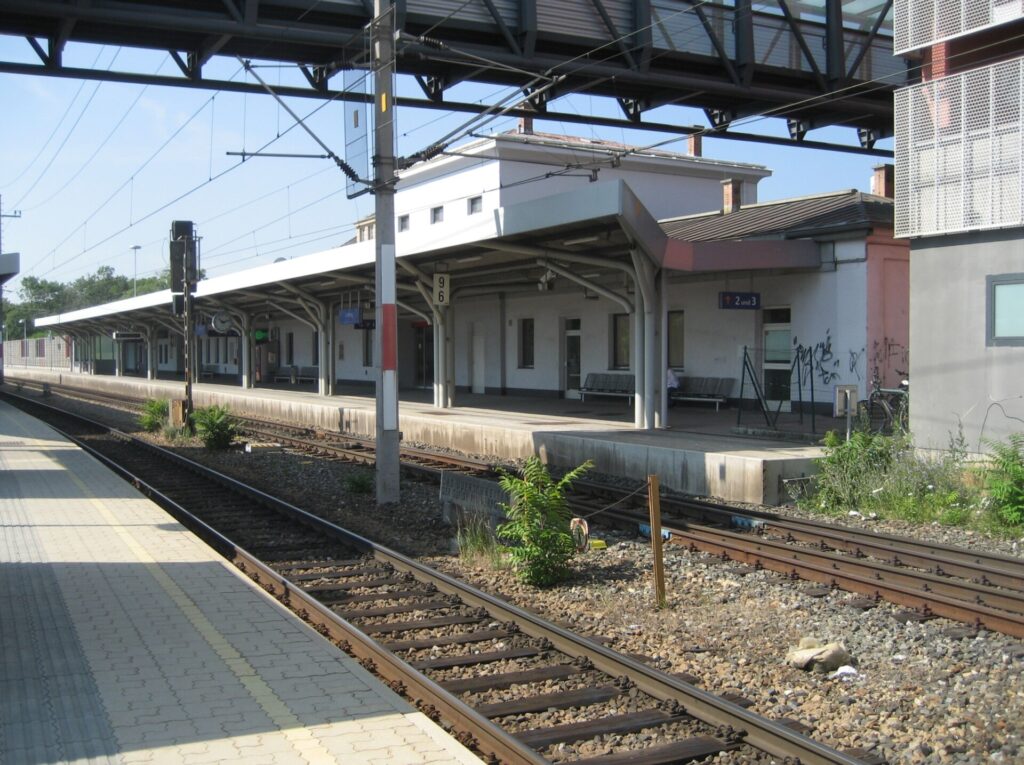 Das weiß verputzte Bahnhofsgebäude von Liesing von der Bahnsteigseite, mit überdachtem Bahnsteig, Gleisen im Vordergrund und einer Fußgängerbrücke darüber, bei blauem Himmel