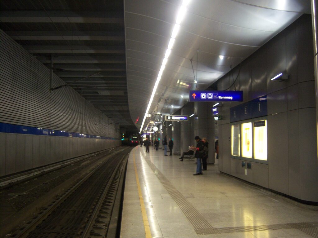 Unterirdischer Bahnsteig der S-Bahn-Station Wien Rennweg mit hellem Steinboden, gewölbter Metalldecke mit Lichtband und blauen Wegweisern neben dem Gleis.