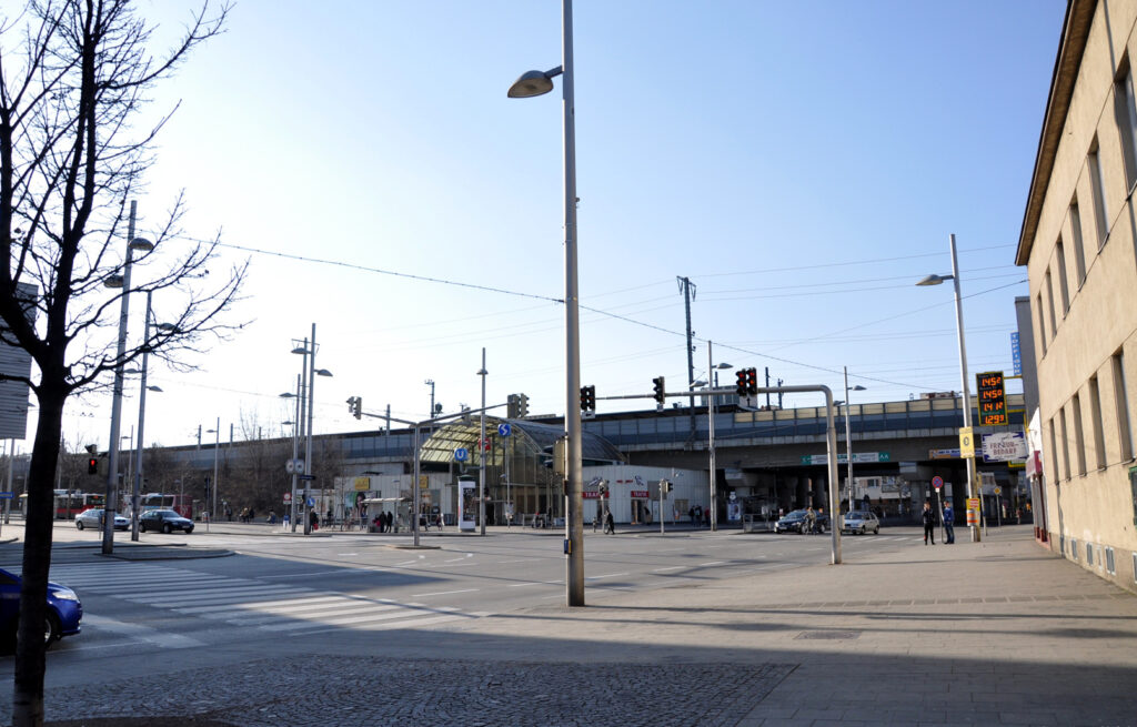 Blick über eine Kreuzung auf die Station Simmering mit gewölbter Glashalle unter der hochliegenden Bahntrasse, davor Ampeln, Straßenlaternen und einige Autos