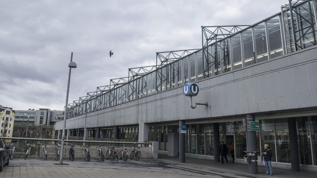 Die obere Ebene der Station Spittelau mit langem Glasaufbau und Stahlfachwerk über einem Betonsockel, daran ein U-Bahn-Würfel, davor Fahrradbügel mit abgestellten Rädern unter bewölktem Himmel