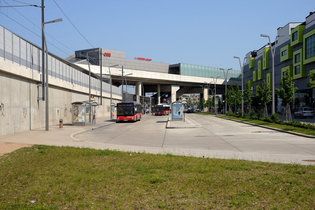Busbahnhof mit zwei roten Bussen vor dem aufgeständerten grauen Stationsgebäude mit ÖBB- und Wiener-Linien-Schriftzug, links eine Lärmschutzwand der Bahn, rechts ein Wohnbau