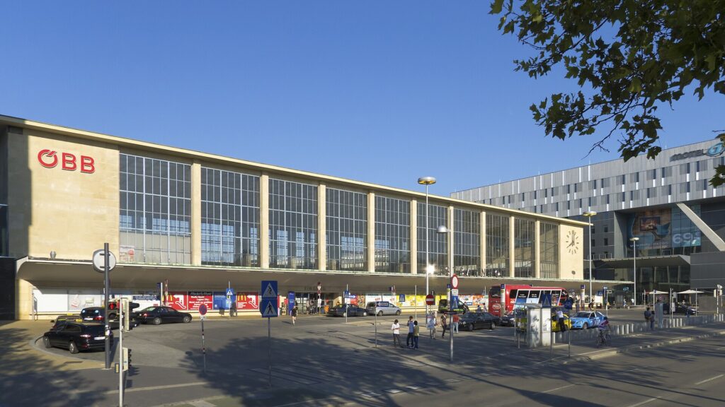 Der Westbahnhof vom Europaplatz aus: die lange, hohe Glasfassade der Bahnhofshalle von 1951 mit dem ÖBB-Logo links und einer Uhr rechts, davor ein Vordach, Taxis und Fußgänger, rechts ein Neubau mit Hotel