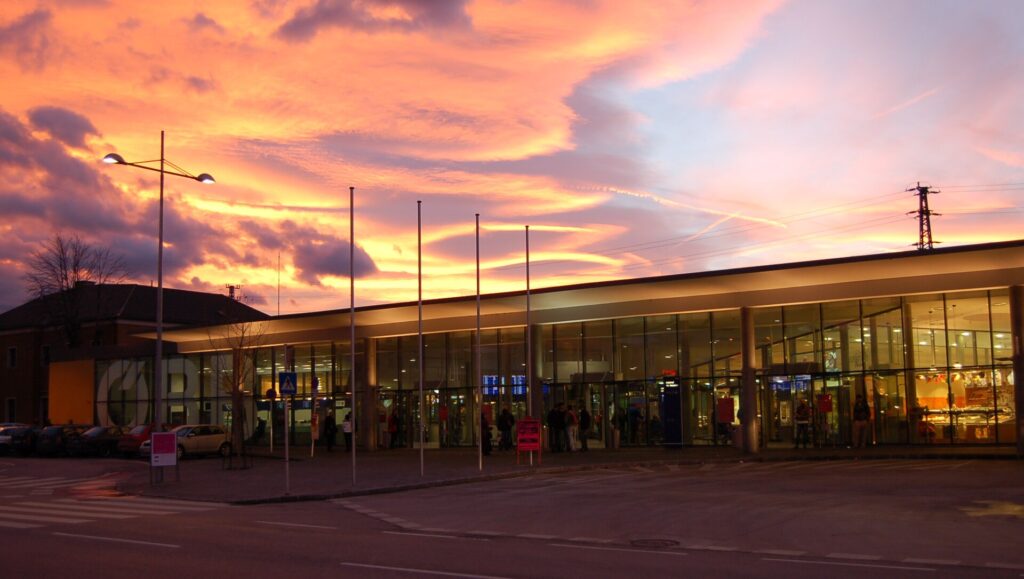 Die verglaste, beleuchtete Bahnhofshalle von Wiener Neustadt mit weit auskragendem Flachdach in der Abenddämmerung, davor Fahnenmasten und die Straße, rosa-oranger Wolkenhimmel