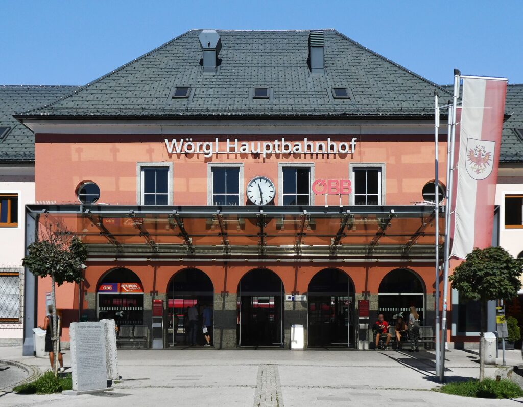 Frontalansicht des lachsfarbenen Mittelbaus mit dem Schriftzug „Wörgl Hauptbahnhof“, Uhr, ÖBB-Logo, gläsernem Vordach und fünf Rundbogenöffnungen unter einem dunklen Walmdach, davor der gepflasterte Vorplatz mit Gedenkstein und Fahnenmast