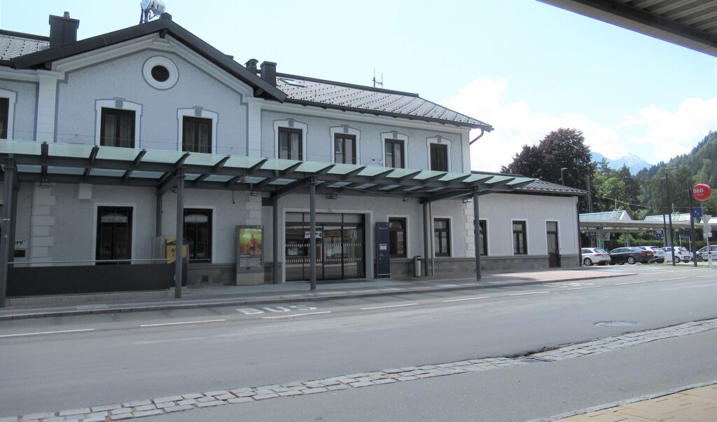 Straßenansicht des hellgrauen, zweigeschoßigen Aufnahmsgebäudes von Zell am See mit Rundfenster im Giebel und langem Glasvordach über dem Eingang, davor eine Busspur mit der Aufschrift BUS, rechts parkende Autos und ein bewaldeter Berghang